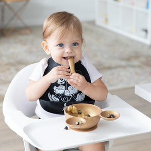 A smiling child in a high chair eats berries and bananas from Bumkins Silicone First Feeding Set: Minnie Mouse Gold.