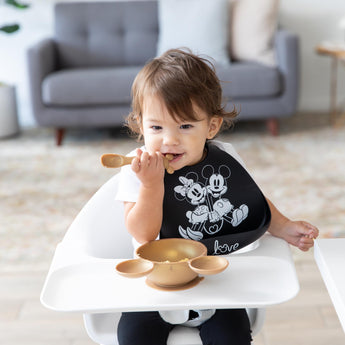 A child sits in a high chair wearing a black bib as they eat a meal from Bumkins Silicone First Feeding Set: Minnie Mouse Gold.