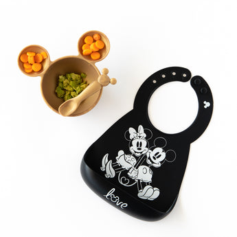 A bib from Bumkins Silicone Bib 2 Pack: Disney Mickey and Minnie Mouse lays next to a gold Mickey-shaped bowl & spoon with food on white.