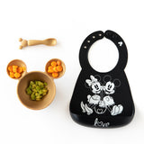 Bumkins Silicone First Feeding Set: Minnie Mouse Gold with carrots and avocado lays neatly next to a black Mickey & Minnie themed bib.