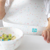 Close-up of a child in a high chair wearing a Bumkins Silicone Bib 2 Pack: Vanilla Sprinkle and Rainbows with a matching bowl nearby.