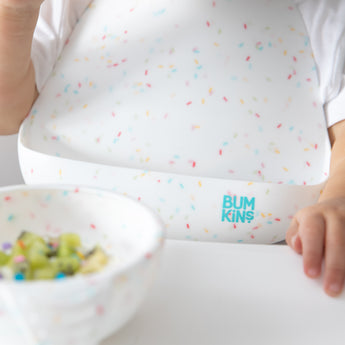 Close-up of a child in a high chair wearing a Bumkins Silicone Bib 2 Pack: Vanilla Sprinkle and Rainbows with a matching bowl nearby.