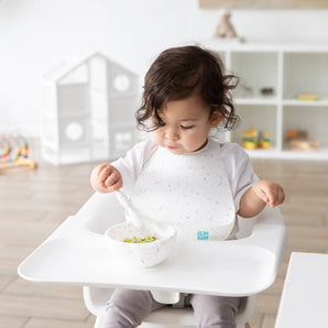 A toddler in high chair wears a Bumkins Silicone Bib 2 Pack: Vanilla Sprinkle and Rainbows while eating from a matching bowl with a spoon.