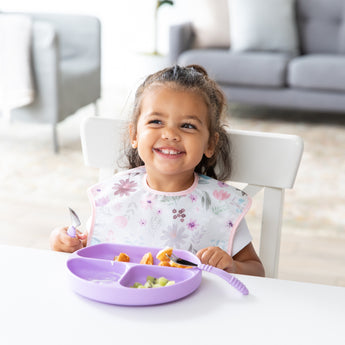 A smiling girl in a floral bib uses Bumkins Spoon + Fork: Lavender to eat from a matching plate at a white table.