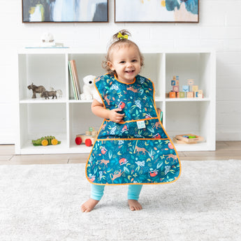 A girl wearing a Bumkins Super-Sized SuperBib®: Jungle smiles in a bright playroom, standing on a cream rug in front of toy-filled shelves.