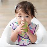 A girl in a floral bib sits in a high chair, drinking from a Bumkins Silicone Starter Cup: Sage.