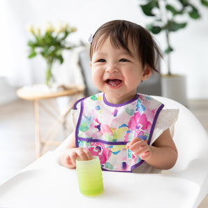 A smiling girl in a high chair wearing a floral bib holds a Bumkins Silicone Starter Cup: Sage in a bright room with plants.