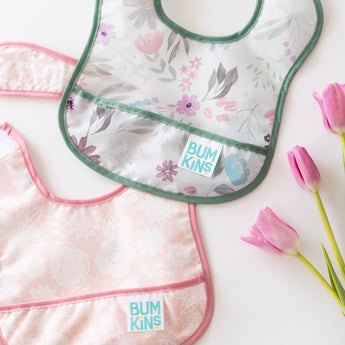 Bumkins Starter Bib 2 Pack: Floral & Lace  rests overlapping on a white background next to three pink tulips.