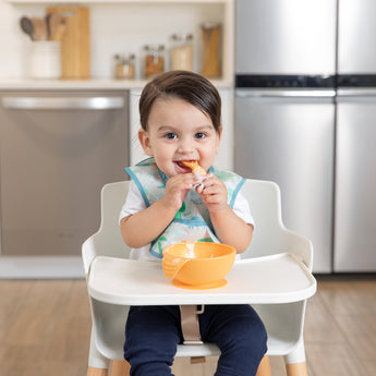 A dark-haired baby in a Bumkins Starter Bib 2 Pack: Dinosaurs & Blue Tropic smiles in a highchair with an orange bowl; kitchen behind.