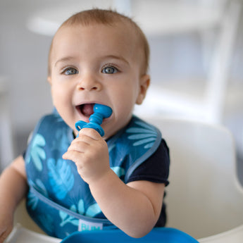 A baby with bright eyes in a blue bib chews a Bumkins Silicone Chewtensils®: Dark Blue.