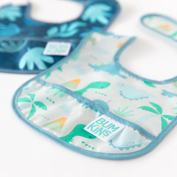 Close-up of Bumkins Starter Bib 2 Pack: Dinosaurs & Blue Tropic. One bib features colorful dinosaurs, palm trees, and a BUMKINS label; the other has a teal design with tropical leaves. Both are waterproof with light blue trim for easy cleaning.