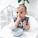 A baby girl in a black & white bib and bow chews a Bumkins Silicone Dipping Spoons 3 Pack: Taffy near a gray bowl on a high chair tray.