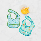 Bumkins Starter Bib 2 Pack: Ocean Life & Whale Tail lays flat on a marble background next to a yellow bowl & spoon set.