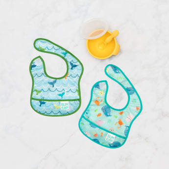 Bumkins Starter Bib 2 Pack: Ocean Life & Whale Tail lays flat on a marble background next to a yellow bowl & spoon set.