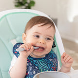A boy wearing a bib from Bumkins Starter Bib 2 Pack: Jungle & Animal Prints in a high chair chews a gray spoon near a gray bowl.