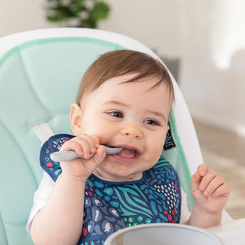 A boy wearing a bib from Bumkins Starter Bib 2 Pack: Jungle & Animal Prints in a high chair chews a gray spoon near a gray bowl.
