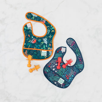 Bumkins Silicone Chewtensils®: Red rests on top of a animal print bib on a marble surface next to another similar bib and orange utensils.