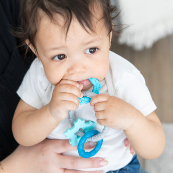 Close-up of a child chewing a Bumkins Silicone Teething Charms: Blue while being held by adult hands.