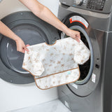A person holds Bumkins Sleeved Bib: Palm Daze in front of an open front-loading washing machine.
