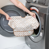 A person holds Bumkins Sleeved Bib: Sand Check in front of an open front-loading washing machine.