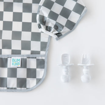 Bumkins Sleeved Bib: Charcoal Check next to baby utensils on a white background, ideal for baby-led weaning.