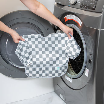 A person holds Bumkins Sleeved Bib: Charcoal Check in front of an open front-loading washing machine.
