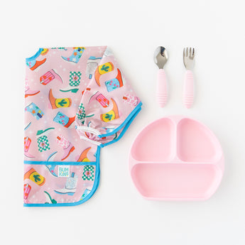 A Bumkins Sleeved Bib 2 Pack: Disco Boots lays folded next to a pink divided plate and matching utensils on a white background.