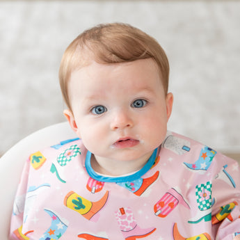 A blue-eyed baby wears a bib from Bumkins On-The-Go Mealtime 6-Piece Set: Boots + Pink.