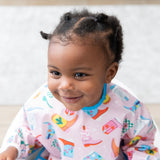 A girl with braided hair smiles, wearing a Bumkins Sleeved Bib: Boots with a blurred beige rug behind.