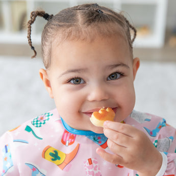 Close-up of a smiling girl in pigtails wearing a Bumkins Sleeved Bib: Boots holding a snack to her mouth.