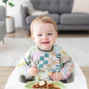 A smiling toddler in a high chair wears a Bumkins Sleeved Bib: Motley Check, holding a spoon with waffles and bananas on a plate in front.