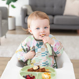 A baby wearing a colorful patterned bib in a high chair eats breakfast foods from a Bumkins Silicone Grip Dish: Sage with matching utensils.