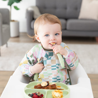 A baby wearing a colorful patterned bib in a high chair eats breakfast foods from a Bumkins Silicone Grip Dish: Sage with matching utensils.