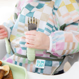 Close-up of a child in a colorful checkered sleeved bib holding Bumkins Spoon + Fork: Sage.