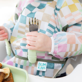 Close-up of a child in a colorful checkered sleeved bib holding Bumkins Spoon + Fork: Sage.