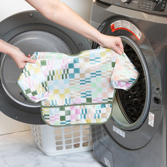 A person holds Bumkins Sleeved Bib: Motley Check in front of an open front-loading washing machine.