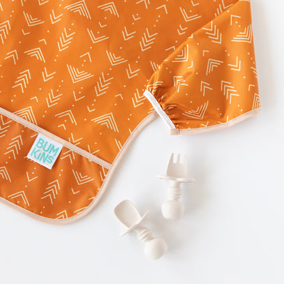 Orange bib with white geometric patterns and a Bumkins label, lying on a white surface. Next to it are a white toddler fork and spoon with textured handles.