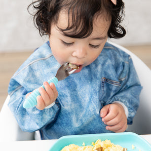 A toddler with curly hair eats scrambled eggs in a high chair, wearing a Bumkins Sleeved Bib: Vintage Denim.