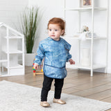 A red-haired child stands in a bright room, wearing a Bumkins Sleeved Bib: Vintage Denim.