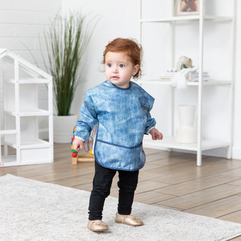 A red-haired child stands in a bright room, wearing a Bumkins Sleeved Bib: Vintage Denim.