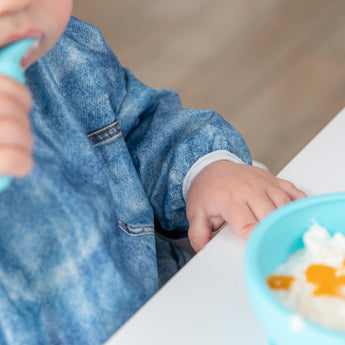 Close-up of a child in a Bumkins Sleeved Bib: Vintage Denim at a white table with a blue bowl of white food; focus is on the elastic cuff.