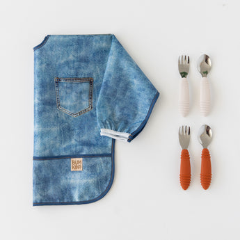 Bumkins Sleeved Bib: Vintage Denim lays folded next to two sets of silicone-handled utensils on a white background.