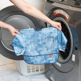 A person holds Bumkins Sleeved Bib: Vintage Denim in front of an open front-loading washing machine.