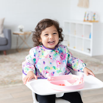 A curly-haired girl smiles in a high chair, wearing a floral bib and eating from a Bumkins Silicone Grip Dish: Pink in a bright playroom.