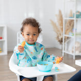 A toddler in a Bumkins Sleeved Bib: Ocean Life sits in a high chair, holding an orange spoon with a blue bowl on the tray.