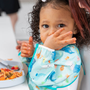 A toddler in a Bumkins Sleeved Bib: Ocean Life eating pasta with sauce all over hands and face.