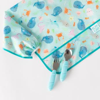 A Bumkins SuperBib® & Sleeved Bib 3-Piece Set in ocean designs, with blue-handled fork and spoon, sits on a white surface.