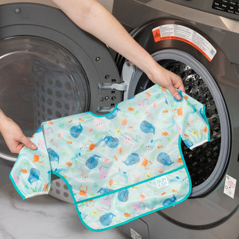 A person holds a bib from Bumkins SuperBib® and Sleeved Bib 3-Piece Set: Rolling With The Waves, Whale Tail, Ocean Life in front of a washer.