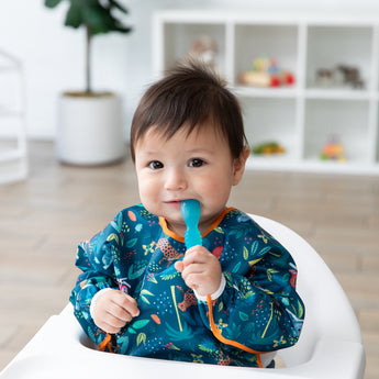 A happy toddler in a Bumkins Sleeved Bib: Jungle holds an blue spoon in their mouth.