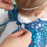 A person adjusts the ribbon closure of a Bumkins Sleeved Bib 2 Pack: Jungle and Animal Print around a child's neck.